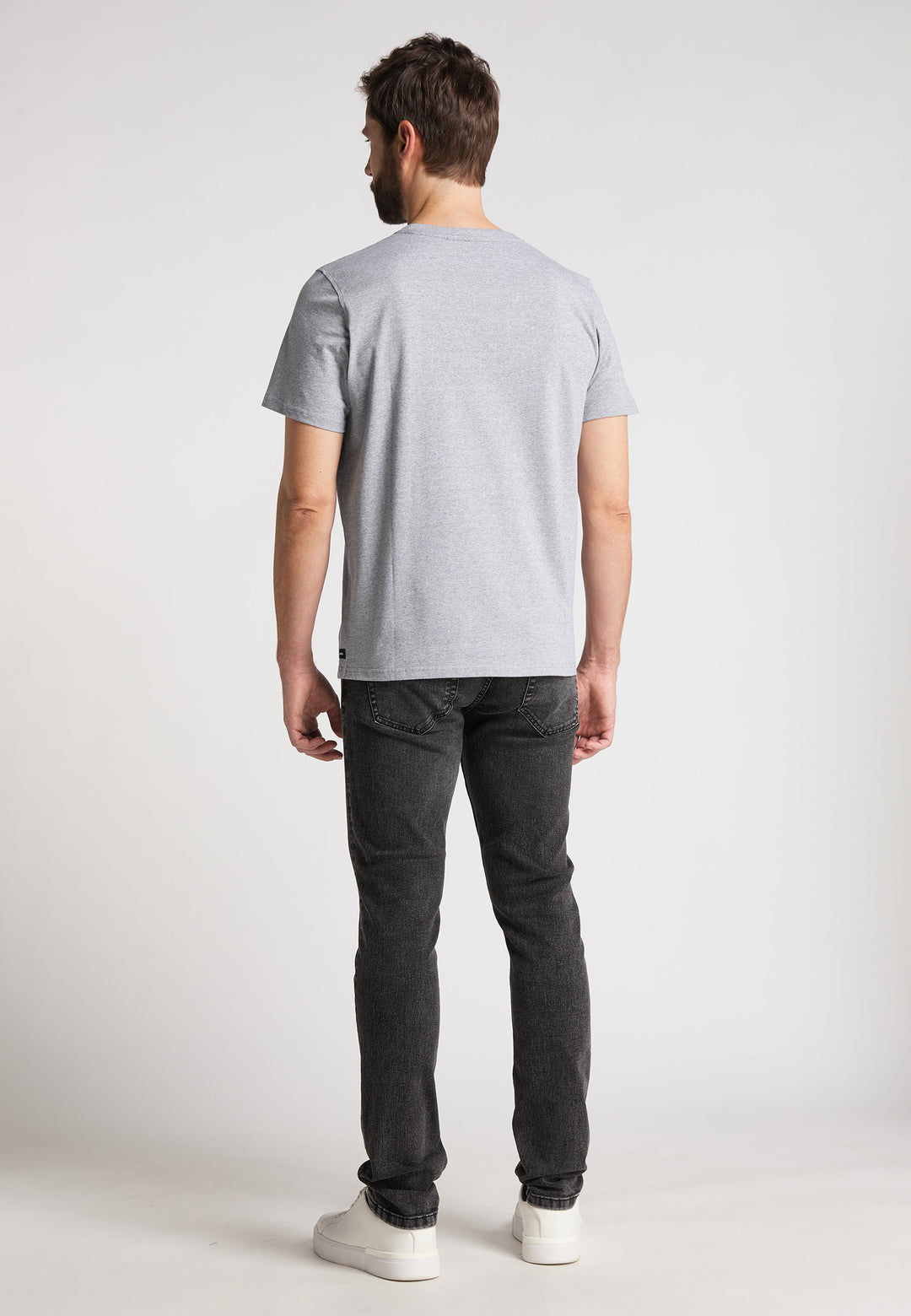 Ein Mann steht mit dem Rücken zur Kamera und trägt das RECARO T-Shirt Originals Embossed aus hellgrauer Baumwolle, dunkelgraue Jeans und weiße Sneakers. Das Shirt hat einen dezenten 3D RECARO Schriftzug auf der Rückseite und einen schlichten hellen Hintergrund.