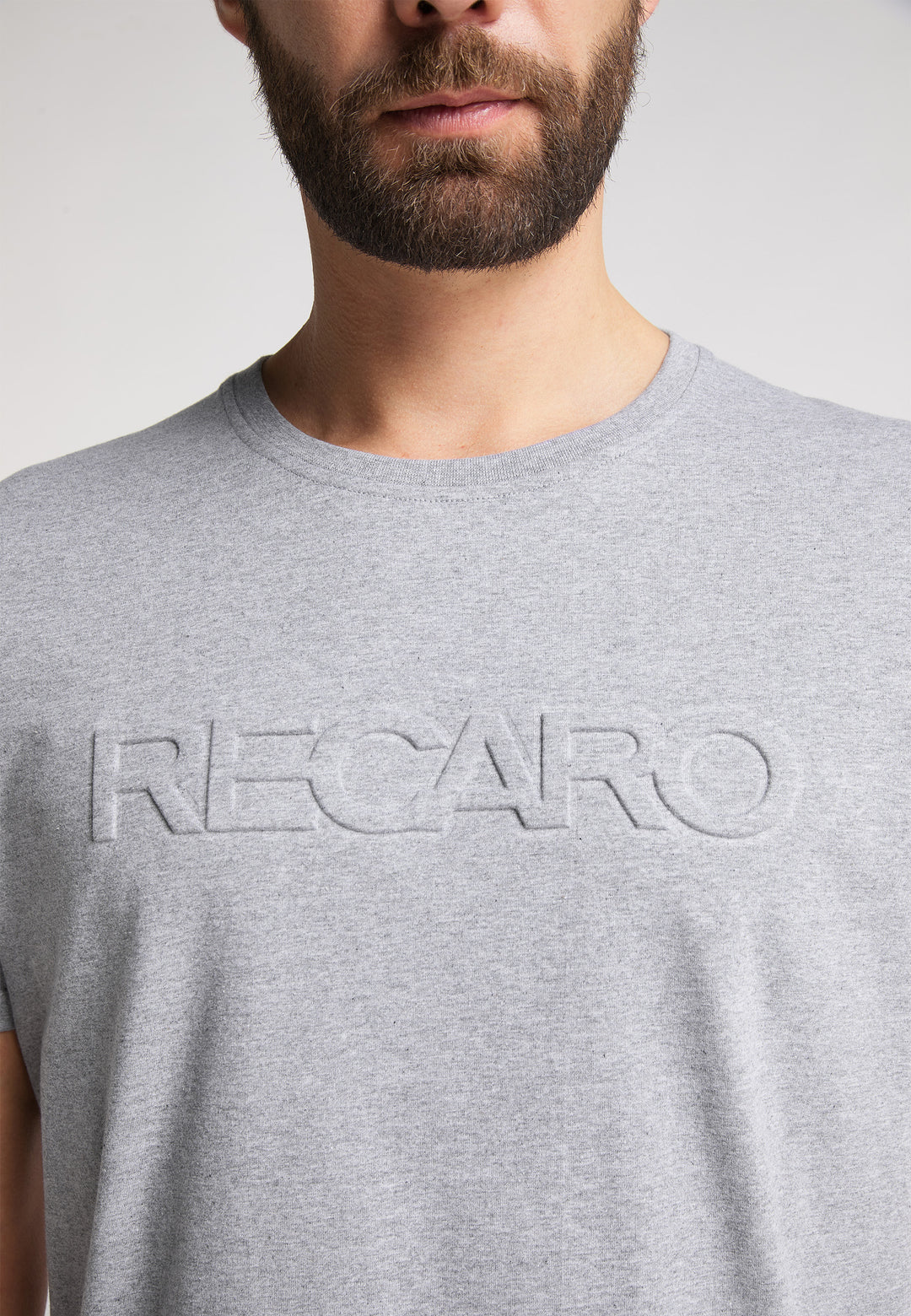 Ein Mann mit gestutztem Bart trägt das RECARO T-Shirt Originals Embossed, ein hellgraues T-Shirt aus 100 % Baumwolle mit einem auffälligen 3D-RECARO-Logo auf der Brust. Auf dem Bild sind nur seine untere Gesichtshälfte und sein Oberkörper zu sehen.