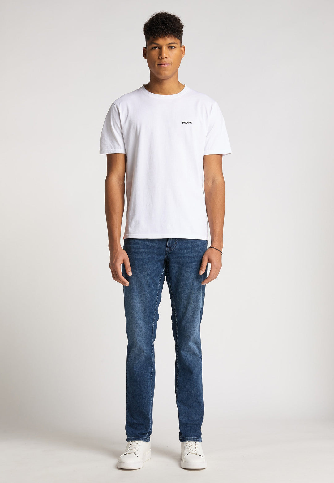 Ein junger Mann blickt nach vorne und trägt ein locker sitzendes Recaro_Merch_Daniel T-Shirt Originals Basic, Bluejeans und weiße Turnschuhe vor einem schlichten hellen Hintergrund.