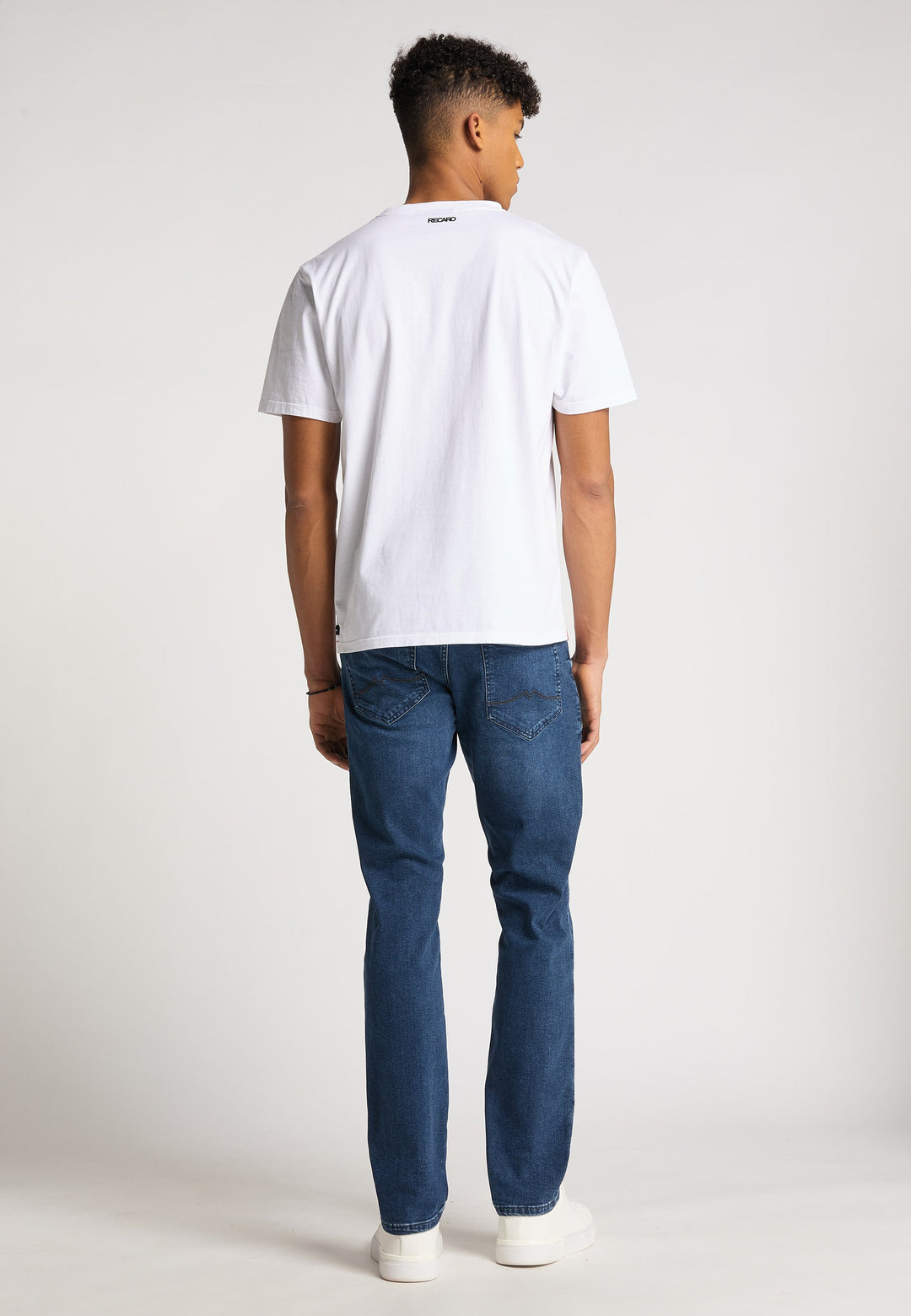 Ein junger Mann steht mit dem Rücken zur Kamera und trägt ein locker sitzendes T-Shirt Originals Basic von Recaro, Bluejeans und weiße Turnschuhe vor einem hellgrauen Studiohintergrund.
