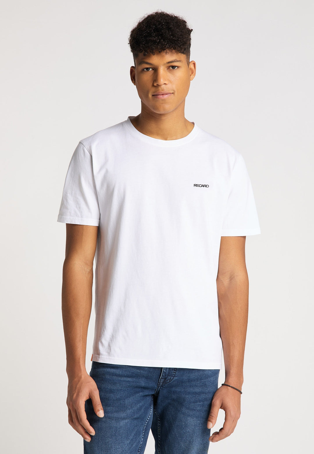 Ein junger Mann mit kurzen lockigen Haaren trägt das Recaro T-Shirt Originals Basic, ein locker sitzendes weißes T-Shirt mit einem kleinen Logo auf der Brust, und Bluejeans, steht vor einem schlichten hellen Hintergrund und blickt in die Kamera.