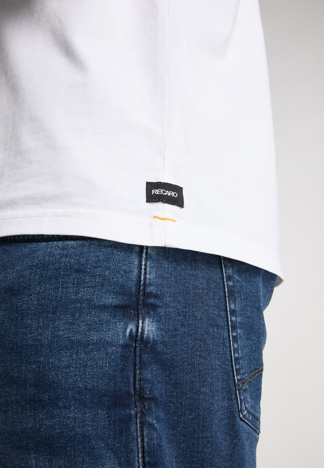 Eine Nahaufnahme einer Person in Blue Jeans und einem weißen Recaro T-Shirt Originals Basic mit einem kleinen Etikett am Saum vor einem schlichten, neutralen Hintergrund.
