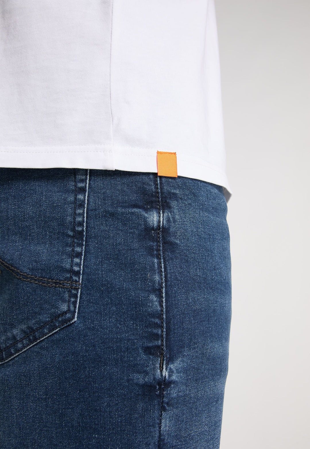 Nahaufnahme einer Person in Blue-Jeans und dem RecaroT-Shirt Originals Basic, einem locker sitzenden weißen T-Shirt mit einem kleinen orangefarbenen Etikett am Saum. Schlichter, neutraler Hintergrund.