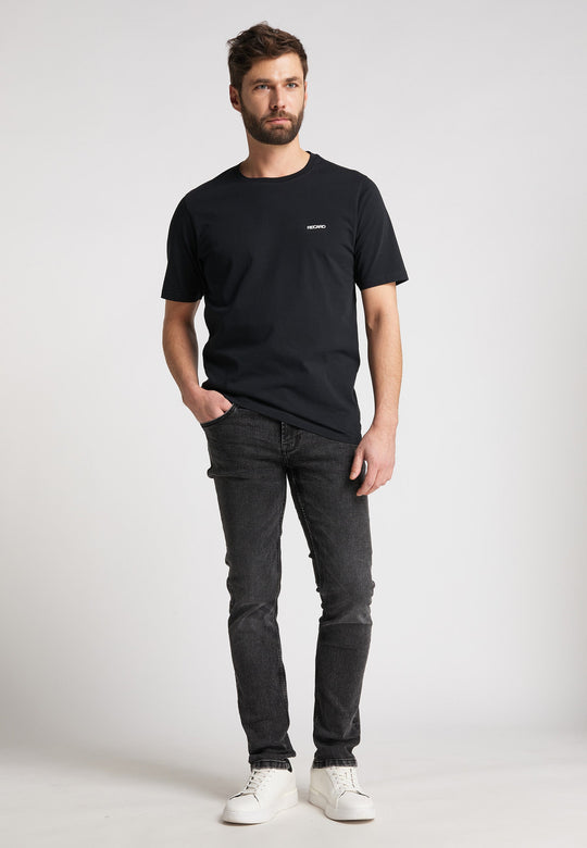 Vor einem weißen Hintergrund steht ein Mann mit kurzen braunen Haaren und Bart. Er trägt ein schwarzes Recaro_Merch_Daniel T-Shirt Originals Basic, dunkle Jeans und weiße Turnschuhe und hat die linke Hand in der Tasche.