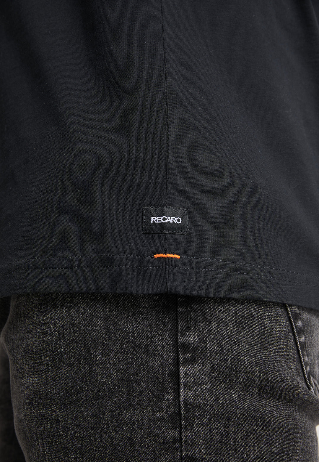 Nahaufnahme des unteren Rückens einer Person, die das T-Shirt Originals Basic von Recaro_Merch_Daniel trägt, ein schwarzes T-Shirt mit lockerer Passform, einem kleinen RECARO-Label und orangefarbenen Nähten, kombiniert mit verwaschenen schwarzen Jeans.