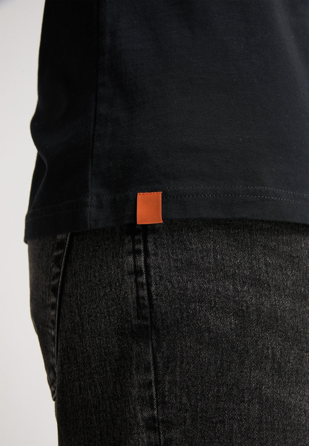 Nahaufnahme einer Person, die das Recaro_Merch_Daniel T-Shirt Originals Basic mit einem orangefarbenen Etikett am Saum trägt, kombiniert mit dunklen Jeans, von der Seite gezeigt.