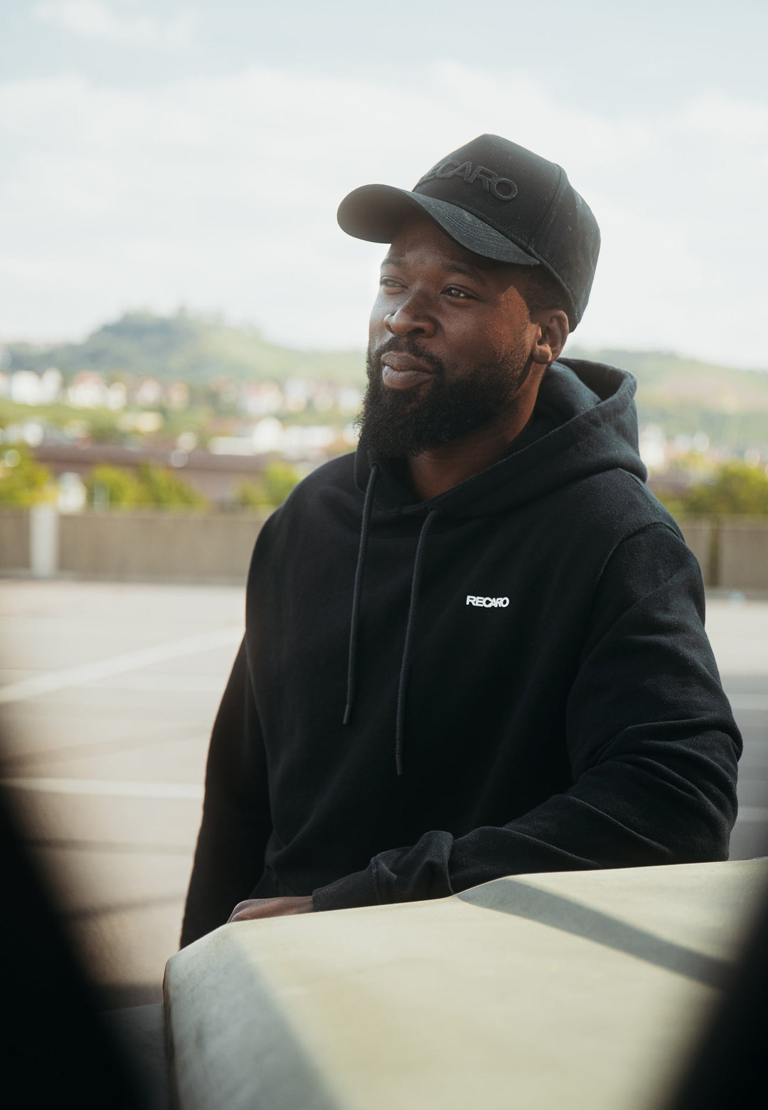 Ein bärtiger Mann in einem locker sitzenden Recaro Hoodie Originals Basic und einer schwarzen Kappe steht im Freien, an eine helle Oberfläche gelehnt, mit einer verschwommenen Stadtlandschaft und viel Grün im Hintergrund.