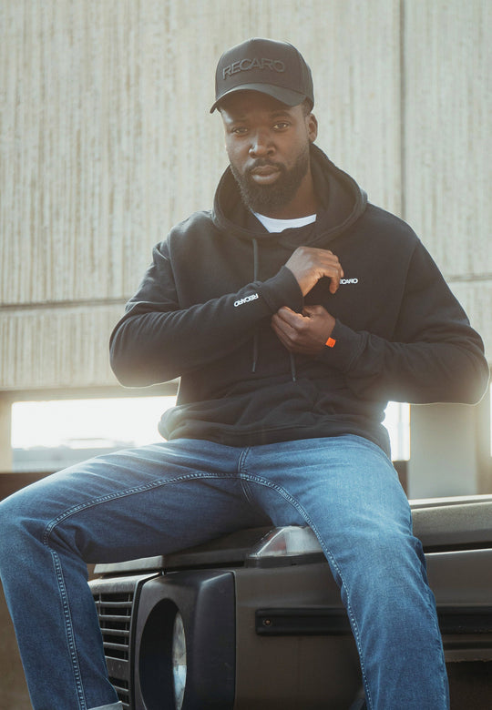 Ein bärtiger Mann in Bluejeans und schwarzer Cap sitzt auf einer Motorhaube. Er trägt einen lässigen Recaro Hoodie Originals Basic aus weichem Baumwollmaterial. Mit neutralem Gesichtsausdruck blickt er in die Kamera und rückt seinen Hoodie zurecht.