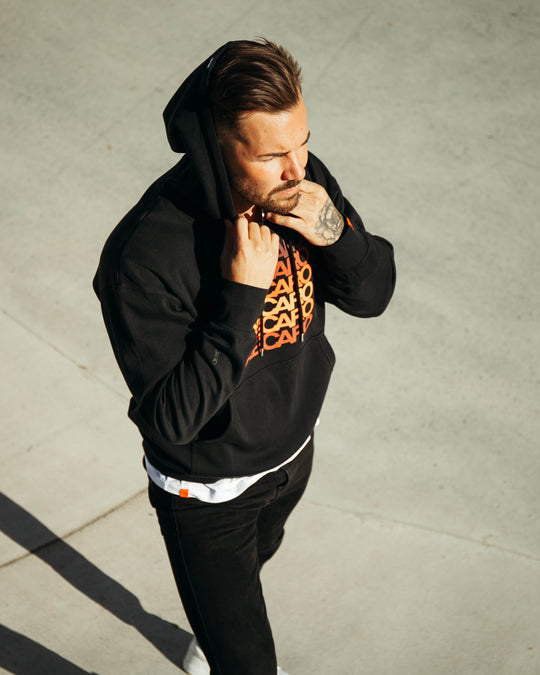 Mit dem RECARO Hoodie Spektrum mit orangefarbenem Text steht ein Mann in schwarzer Hose und weißen Schuhen an einem sonnigen Tag im Freien, hält die Kapuze und blickt mit coolem Retro-Style zur Seite.