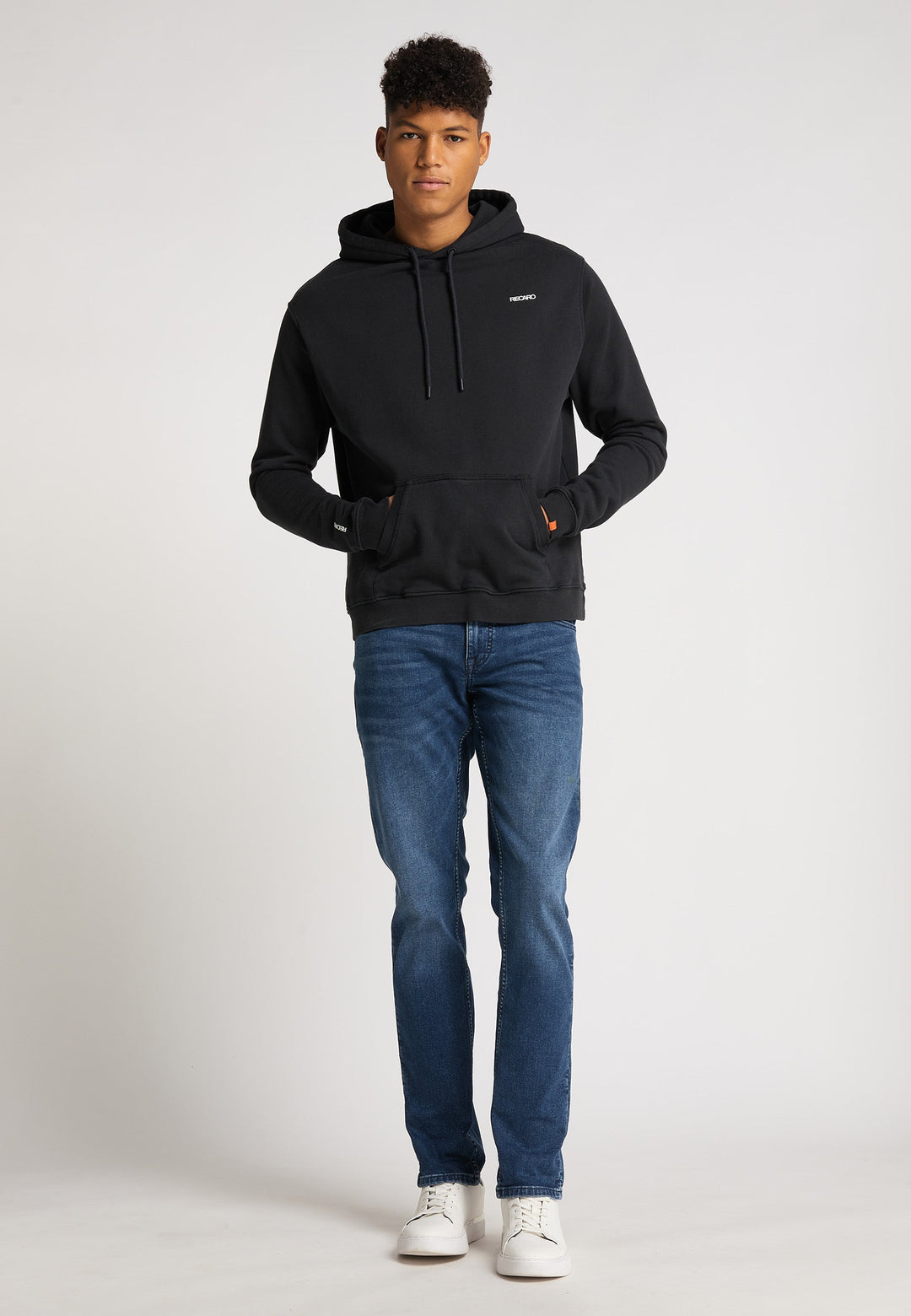 Ein junger Mann steht vor einem schlichten Hintergrund und trägt einen locker sitzenden Hoodie Originals Basic von Recaro_Merch_Daniel, dazu blaue Jeans und weiße Turnschuhe. Er blickt nach vorne, die Hände in den Taschen seines Hoodies.