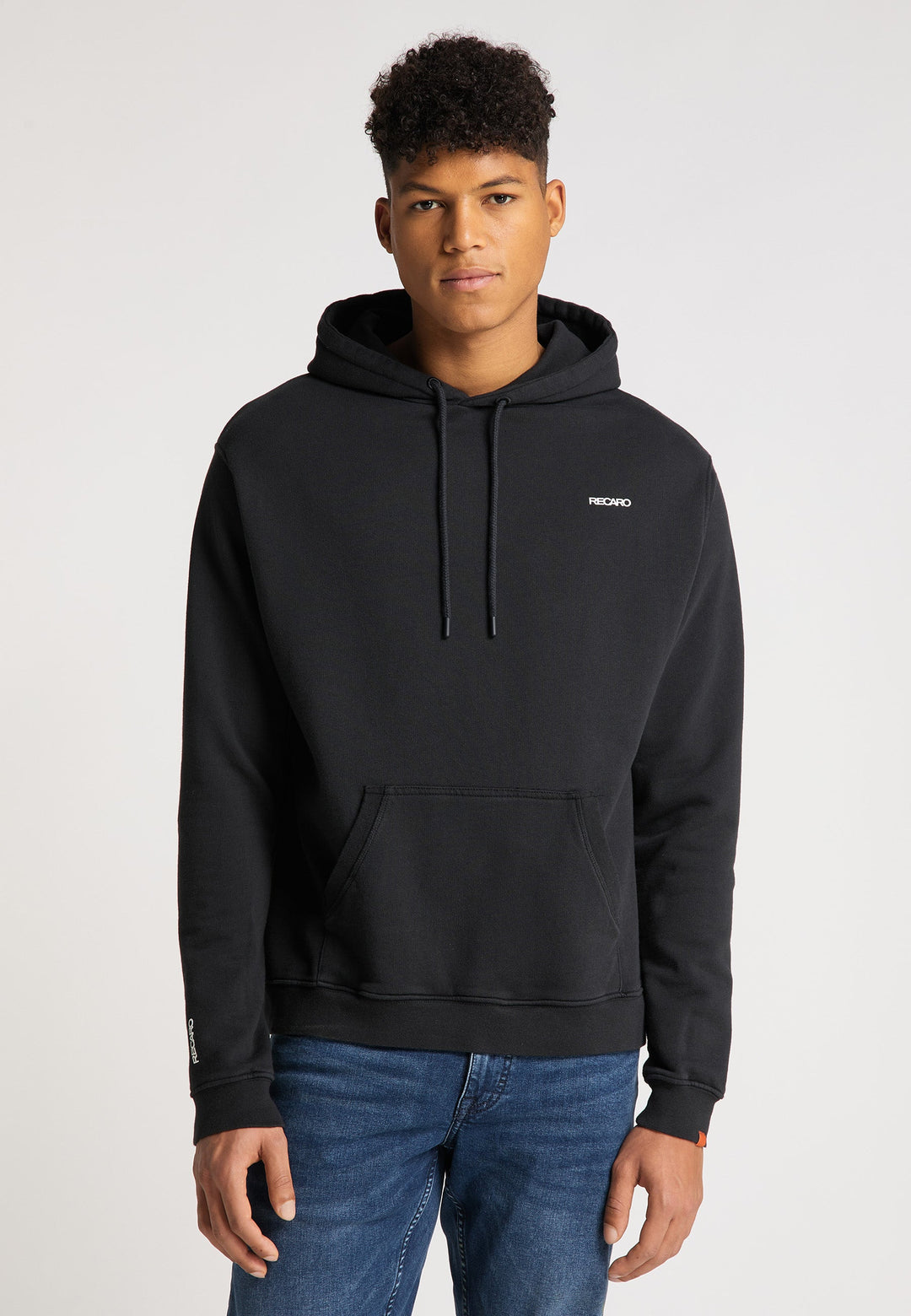 Ein junger Mann mit kurzen lockigen Haaren, der einen schwarzen Recaro_Merch_Daniel Hoodie Originals Basic aus weicher Baumwolle und Blue Jeans trägt, steht vor einem schlichten hellen Hintergrund mit Blick in die Kamera. Der Hoodie trägt ein kleines weißes Logo auf der linken Brust.