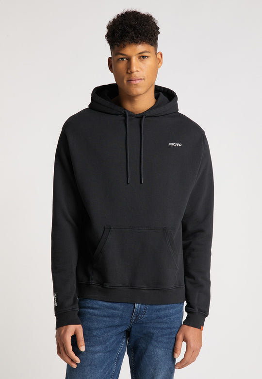 Ein junger Mann mit kurzen lockigen Haaren, der einen schwarzen Recaro_Merch_Daniel Hoodie Originals Basic aus weicher Baumwolle und Blue Jeans trägt, steht vor einem schlichten hellen Hintergrund mit Blick in die Kamera. Der Hoodie trägt ein kleines weißes Logo auf der linken Brust.