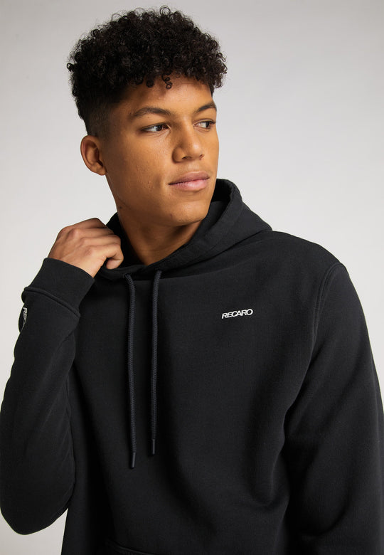 Ein junger Mann mit kurzen lockigen Haaren trägt den lässig geschnittenen Recaro Hoodie Originals Basic aus weicher Baumwolle und blickt vor einem schlichten hellen Hintergrund zur Seite.