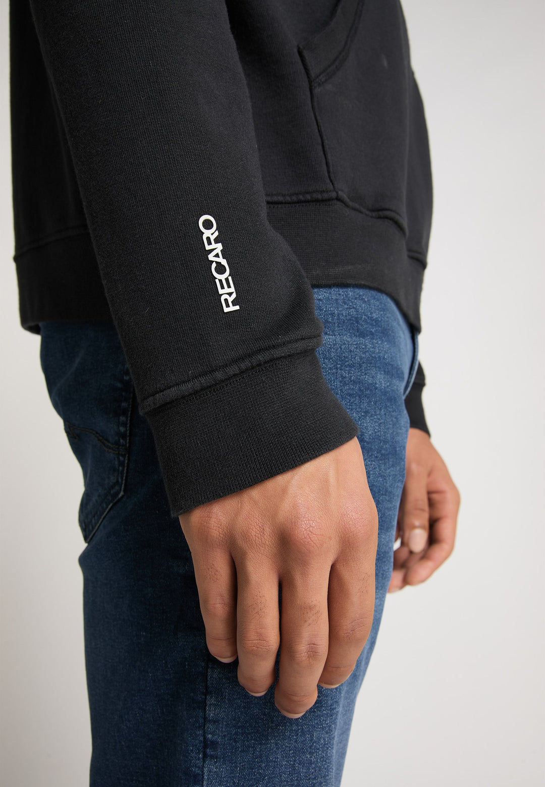 Nahaufnahme des rechten Arms und der rechten Hand einer Person, die den Recaro Hoodie Originals Basic aus schwarzer Baumwolle mit weißem Schriftzug auf dem Ärmel trägt, kombiniert mit Blue Jeans. Die Person steht vor einem schlichten Hintergrund.