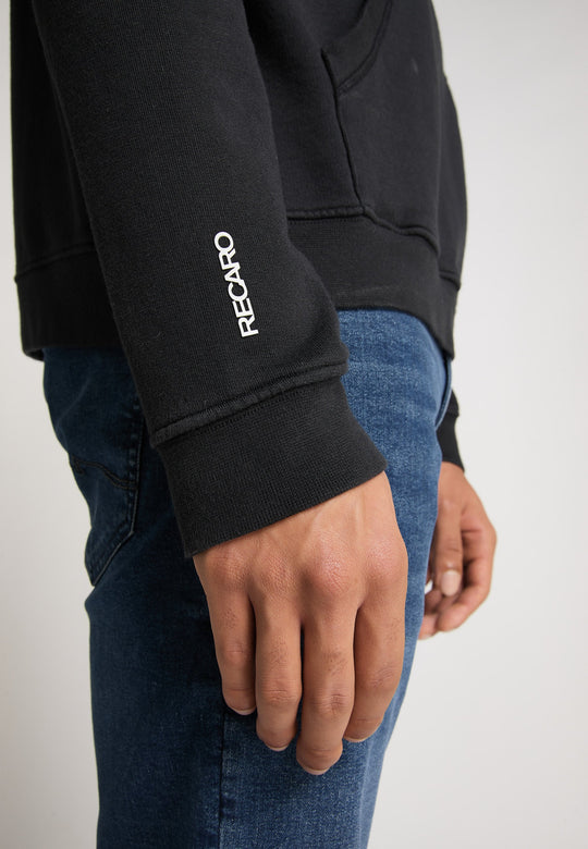 Nahaufnahme des rechten Arms und der rechten Hand einer Person, die den Recaro Hoodie Originals Basic aus schwarzer Baumwolle mit weißem Schriftzug auf dem Ärmel trägt, kombiniert mit Blue Jeans. Die Person steht vor einem schlichten Hintergrund.