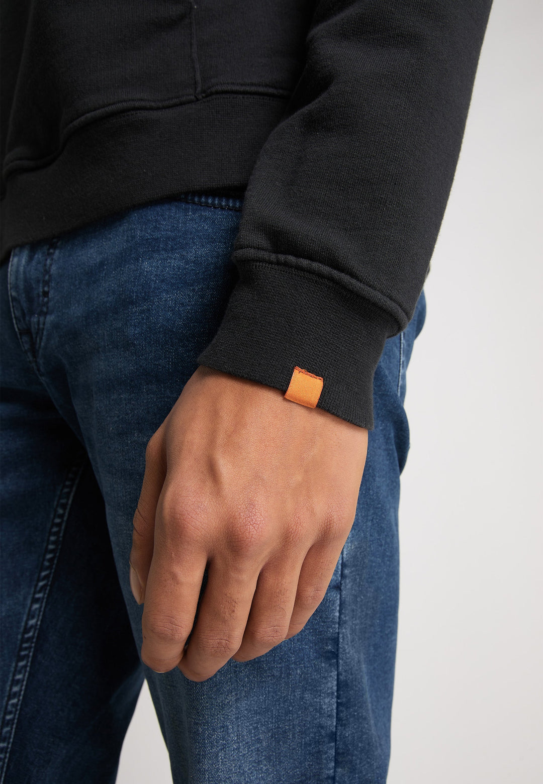 Eine Person trägt einen Recaro Hoodie Originals Basic mit einem orangefarbenen Etikett am Bündchen und Bluejeans und steht mit entspannt herabhängendem Arm da.