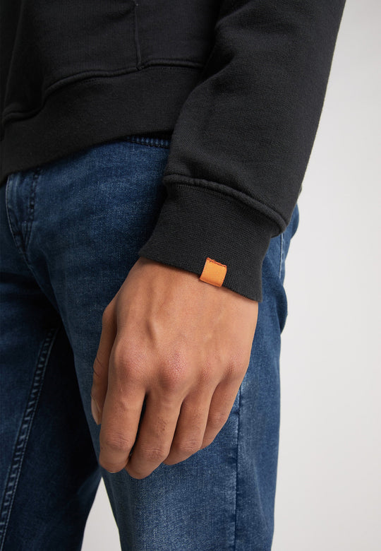 Eine Person trägt einen Recaro Hoodie Originals Basic mit einem orangefarbenen Etikett am Bündchen und Bluejeans und steht mit entspannt herabhängendem Arm da.