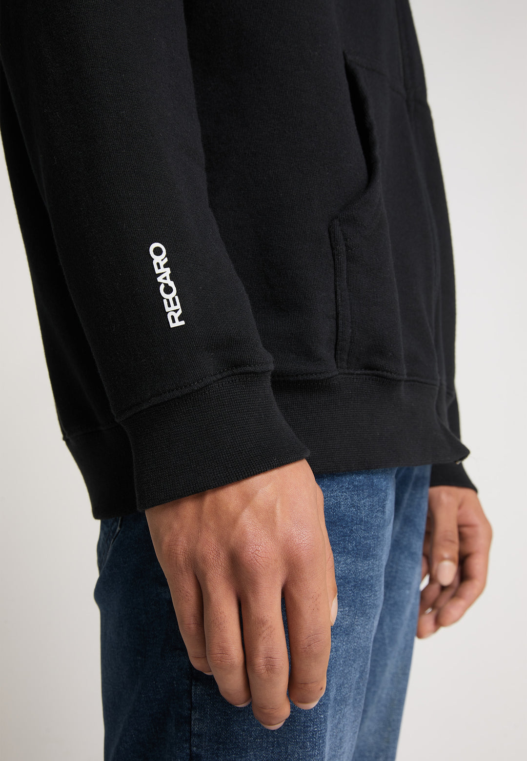 Nahaufnahme einer Person, die die schwarze Hoodiejacke Originals von Recaro trägt, mit einem weißen Logo auf dem Ärmel und kombiniert mit Bluejeans vor einem schlichten, hellen Hintergrund für einen lässigen, entspannten Stil.