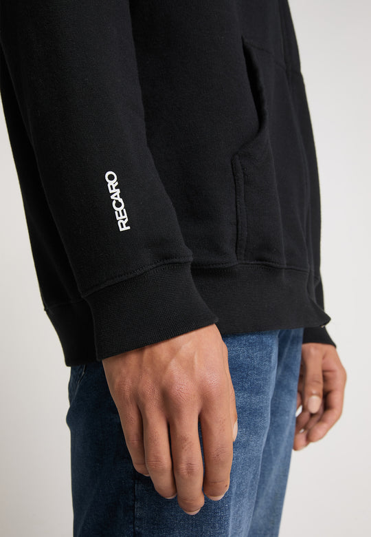 Nahaufnahme einer Person, die die schwarze Hoodiejacke Originals von Recaro trägt, mit einem weißen Logo auf dem Ärmel und kombiniert mit Bluejeans vor einem schlichten, hellen Hintergrund für einen lässigen, entspannten Stil.