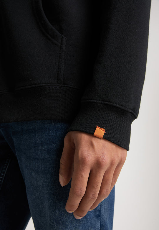 Eine Person in Blue Jeans trägt eine schwarze Recaro Hoodiejacke mit einem kleinen orangefarbenen Etikett am Bündchen, die Hand hängt entspannt an der Seite.