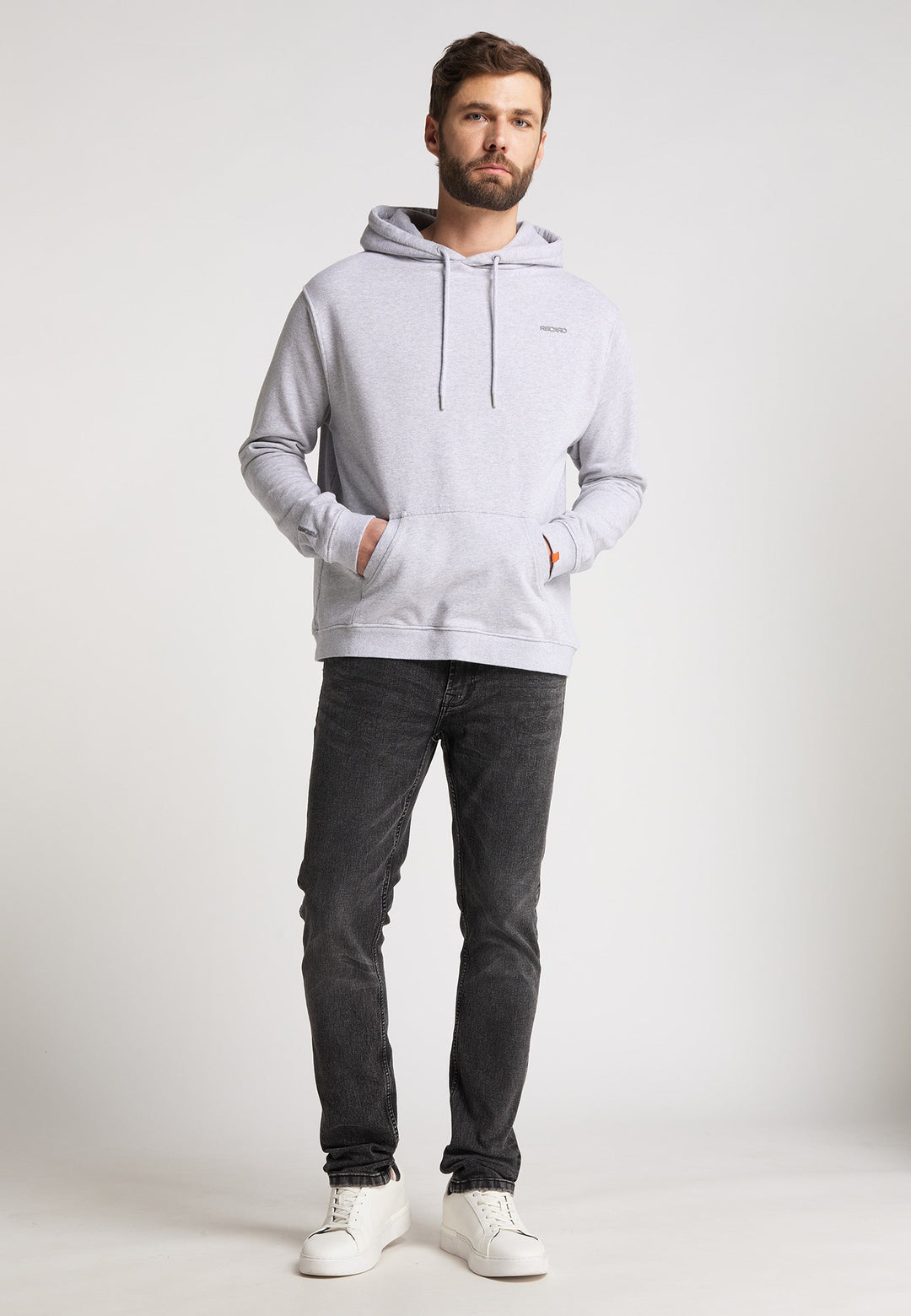 Ein Mann steht mit Blick nach vorne in einem RECARO  Hoodie Originals Basic aus weicher Baumwolle, kombiniert mit schwarzen Jeans und weißen Turnschuhen, die Hände in der Hoodietasche vor einem schlichten hellen Hintergrund.