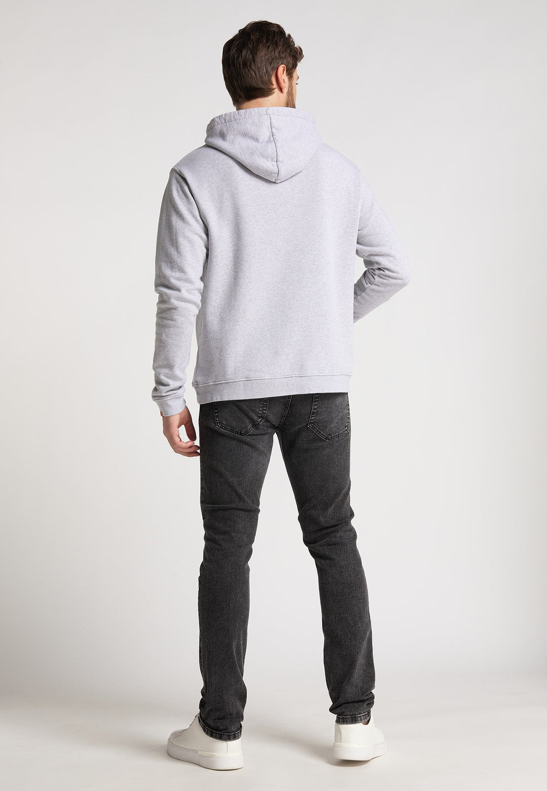 Ein Mann blickt von der Kamera weg und trägt einen hellgrauen Hoodie Originals Basic von RECARO, dunkelgraue Jeans und weiße Turnschuhe vor einem schlichten hellen Hintergrund.