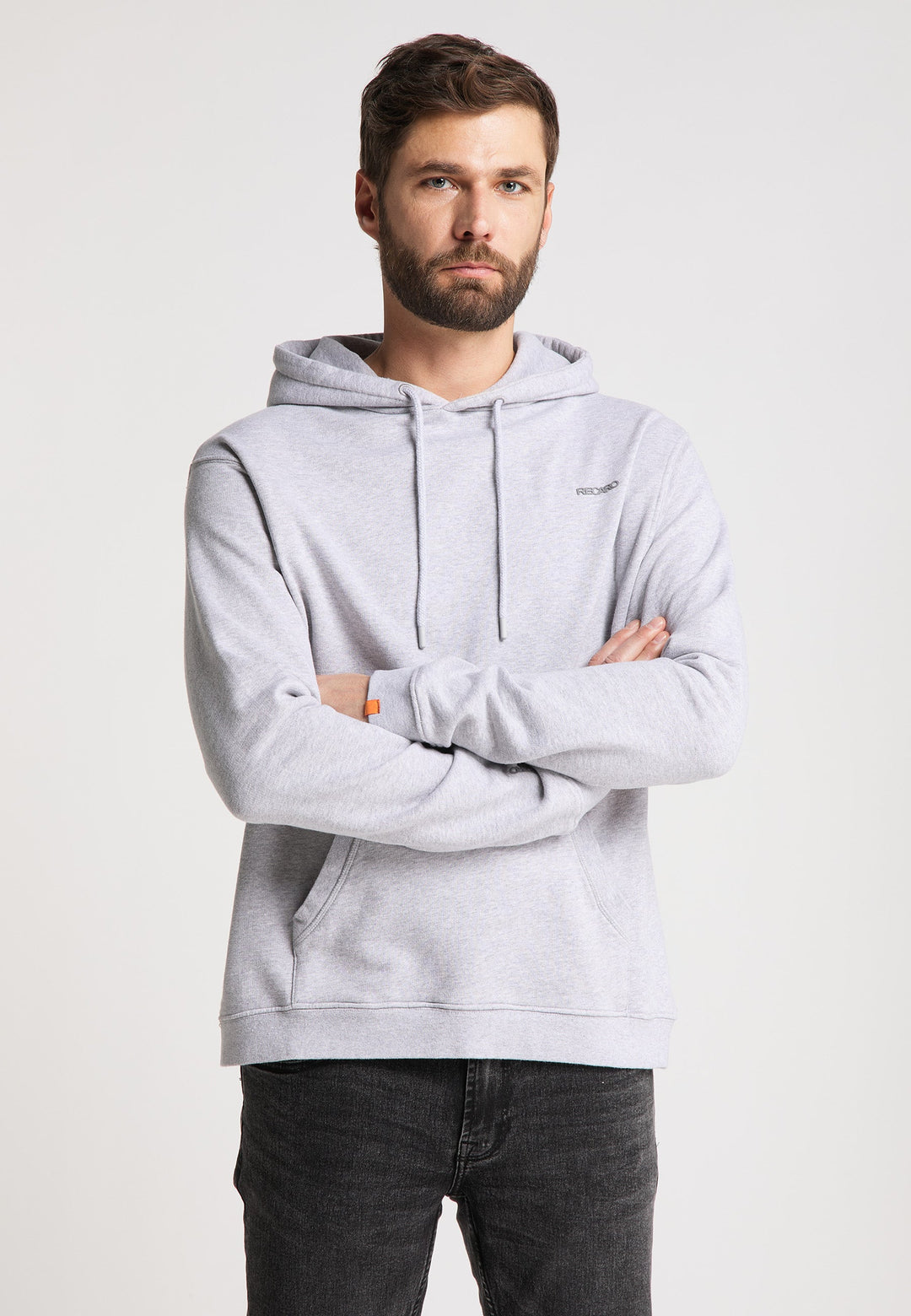 Ein Mann mit Bart und kurzen Haaren steht mit verschränkten Armen nach vorne gerichtet da und trägt einen locker sitzenden Recaro Hoodie Originals Basic und dunkle Jeans vor einem schlichten hellen Hintergrund.