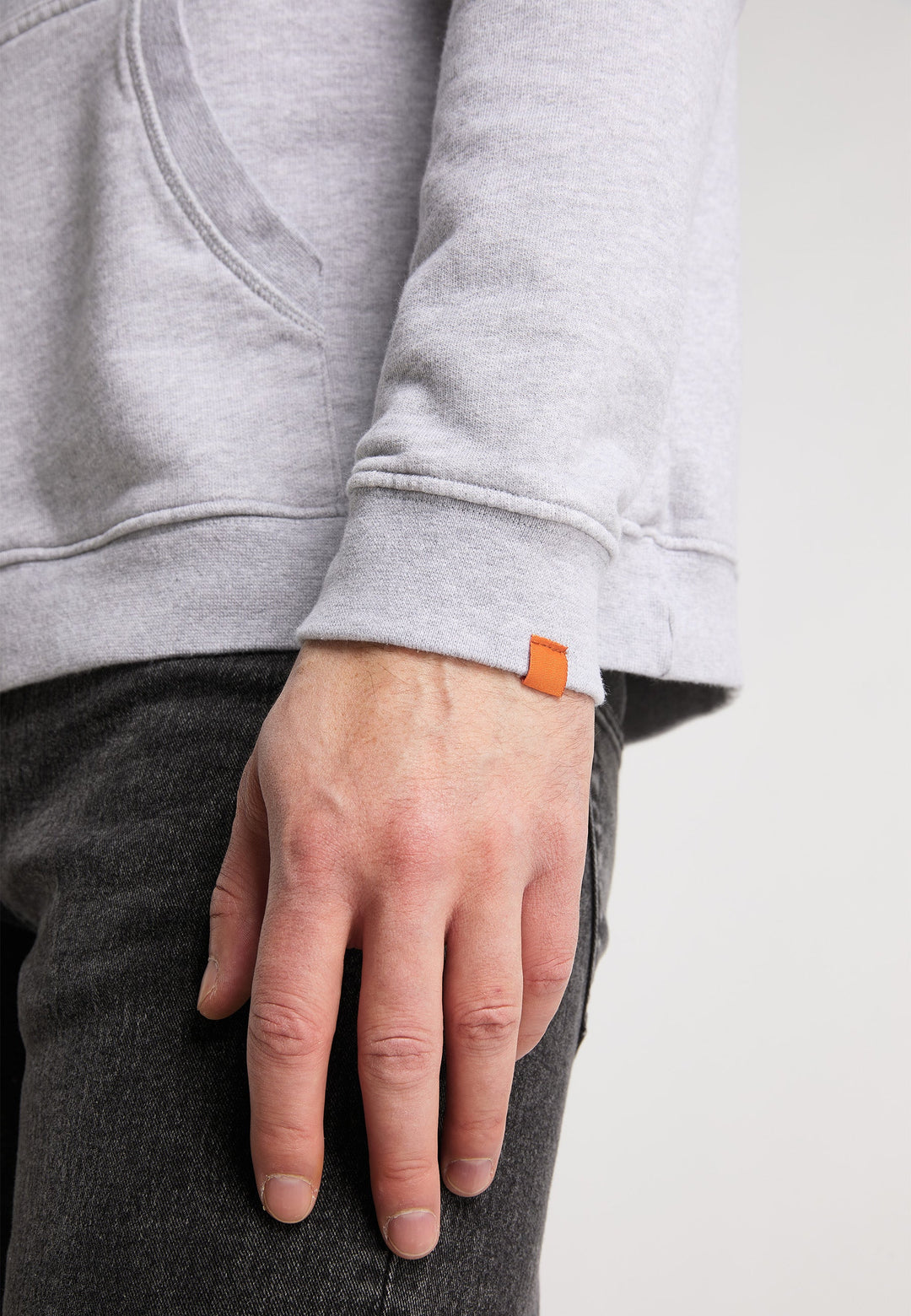 Eine Nahaufnahme der linken Hand einer Person, die an ihrer Seite ruht. Sie trägt einen locker sitzenden hellgrauen Hoodie Originals Basic von Recaro_Merch_Daniel mit einem kleinen orangefarbenen Etikett am Ärmel und dunkelgraue Hosen vor einem schlichten hellen Hintergrund.