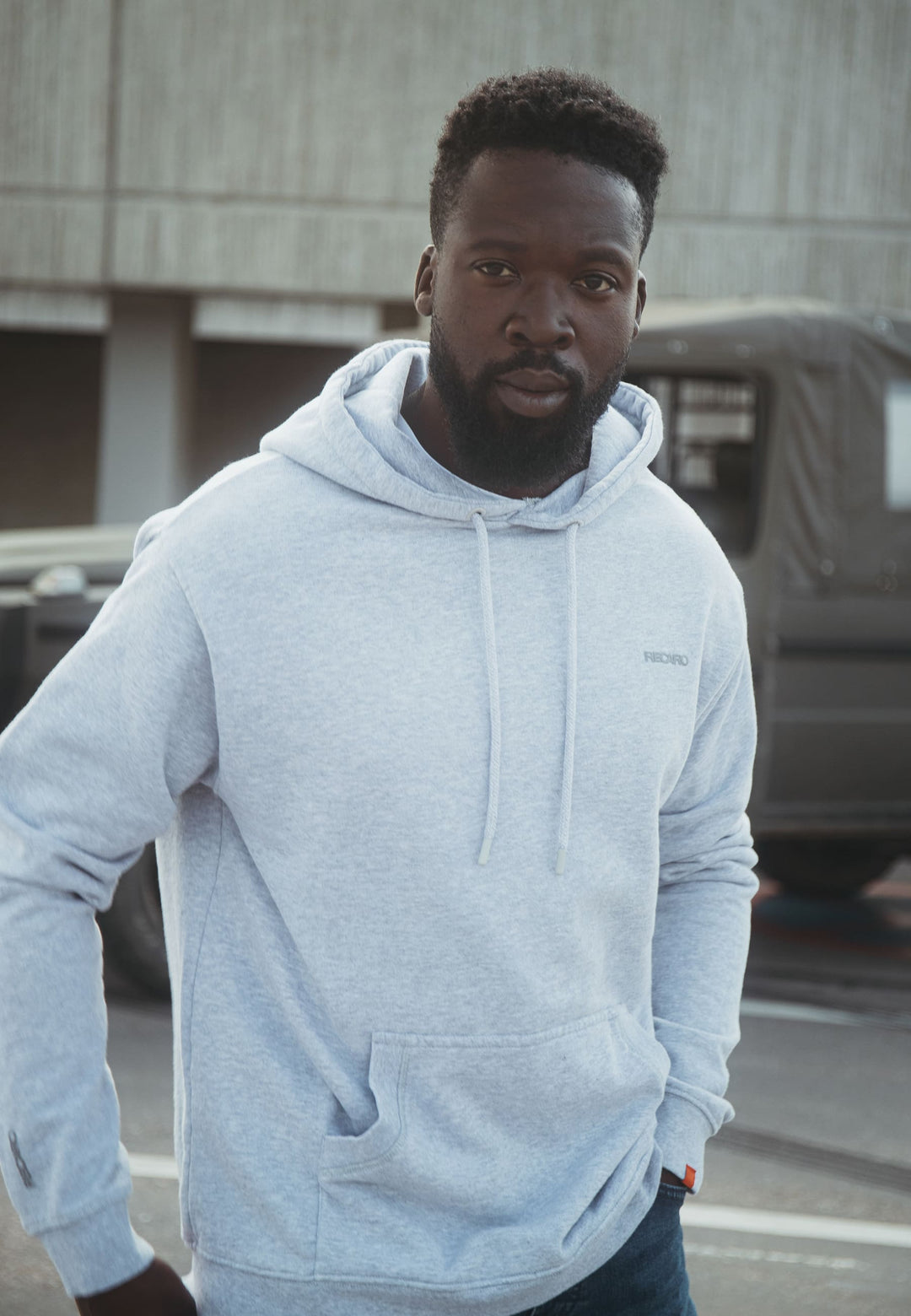 Ein bärtiger Mann in einem locker sitzenden, hellgrauen Recaro_Merch_Daniel Hoodie Originals Basic aus weicher Baumwolle steht im Freien neben einem geparkten Fahrzeug und blickt in die Kamera, im Hintergrund ist ein Betongebäude zu sehen.