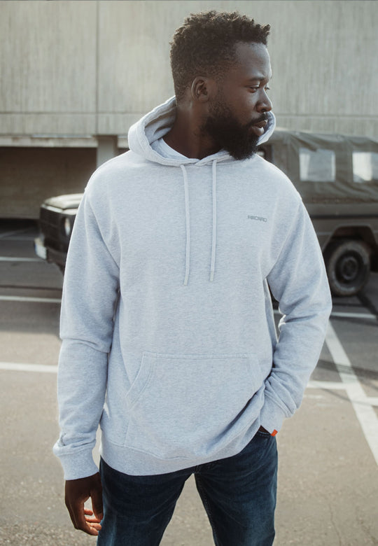 Ein Mann in einem locker sitzenden Recaro_Merch_Daniel Hoodie Originals Basic aus weicher Baumwolle und Bluejeans steht auf einem Parkplatz und blickt mit einer Hand in der Tasche zur Seite. Hinter ihm sind ein dunkles Fahrzeug und ein Betongebäude zu sehen.