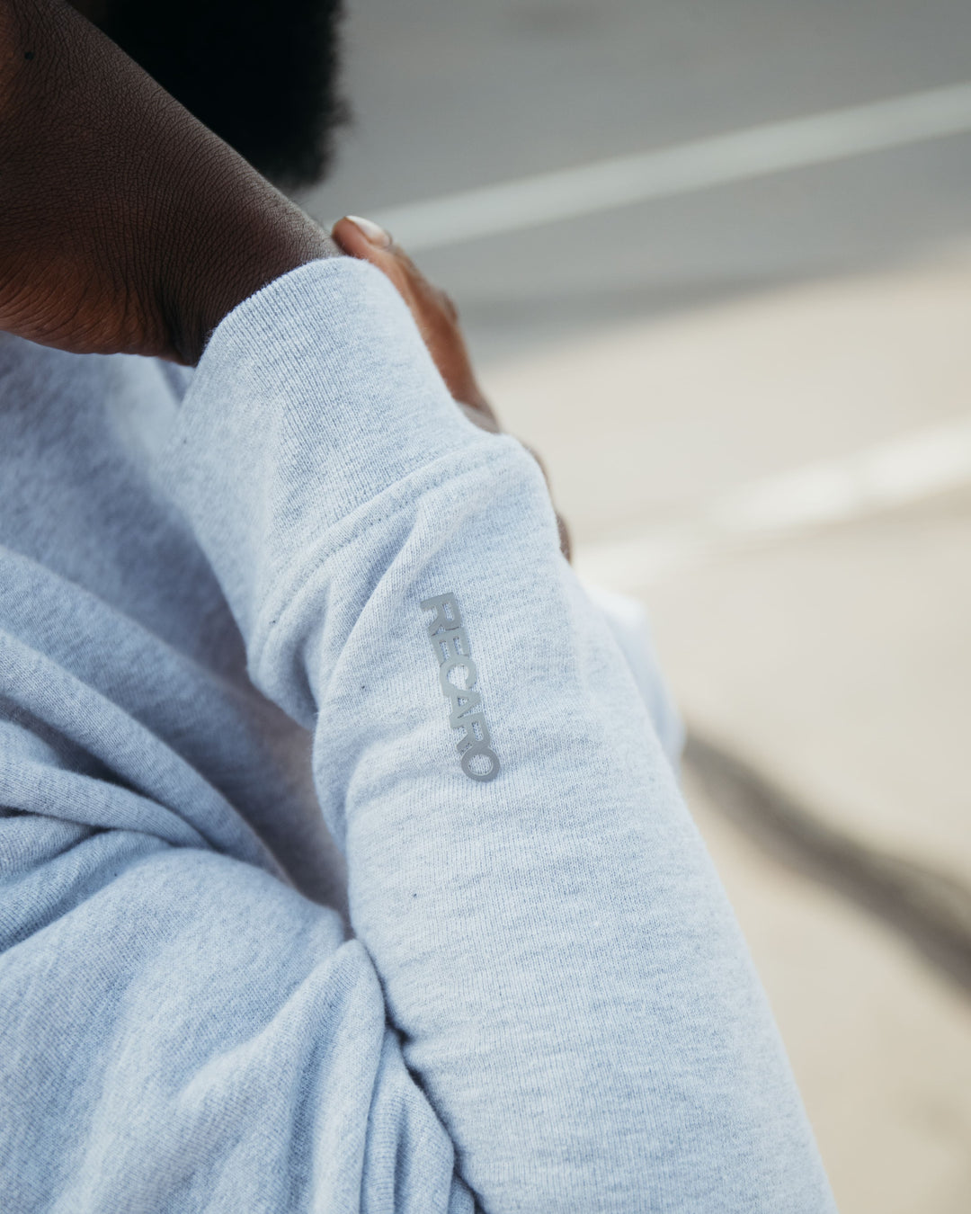 Nahaufnahme einer Person in einem hellgrauen Recaro_Merch_Daniel Hoodie Originals Basic, Relaxed Fit, mit „REBRO“ in kleinen grauen Buchstaben auf dem Ärmel. Die weiche Baumwolle hebt sich vor einem unscharfen Hintergrund mit Straße und Gehweg ab.