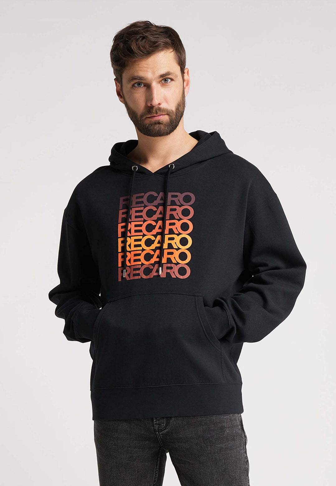 Ein Mann mit kurzen braunen Haaren und Bart trägt den RECARO  Hoodie Spektrum mit dem Schriftzug „RECARO“ in einem Retro-Farbverlauf aus Rot und Orange auf der Brust. Er steht mit einer Hand in der Tasche vor einem schlichten Hintergrund.