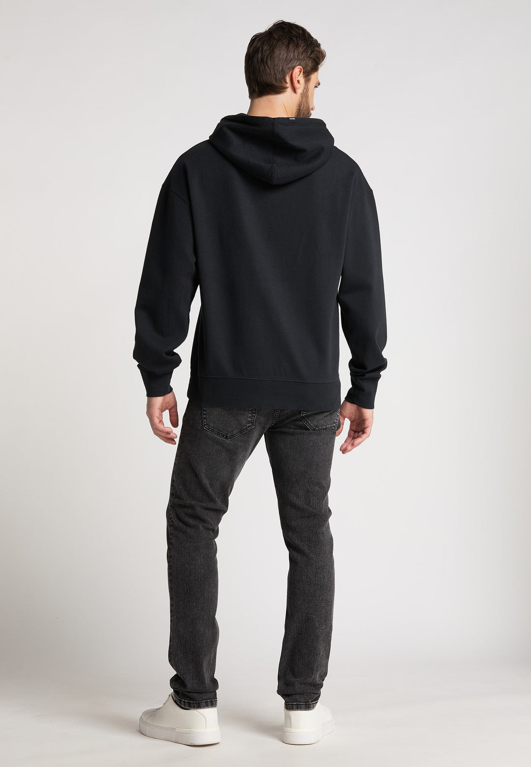 Ein Mann blickt von der Kamera weg und trägt einen RECARO Hoodie Spektrum, dunkle Jeans und weiße Turnschuhe vor einem schlichten hellen Hintergrund.