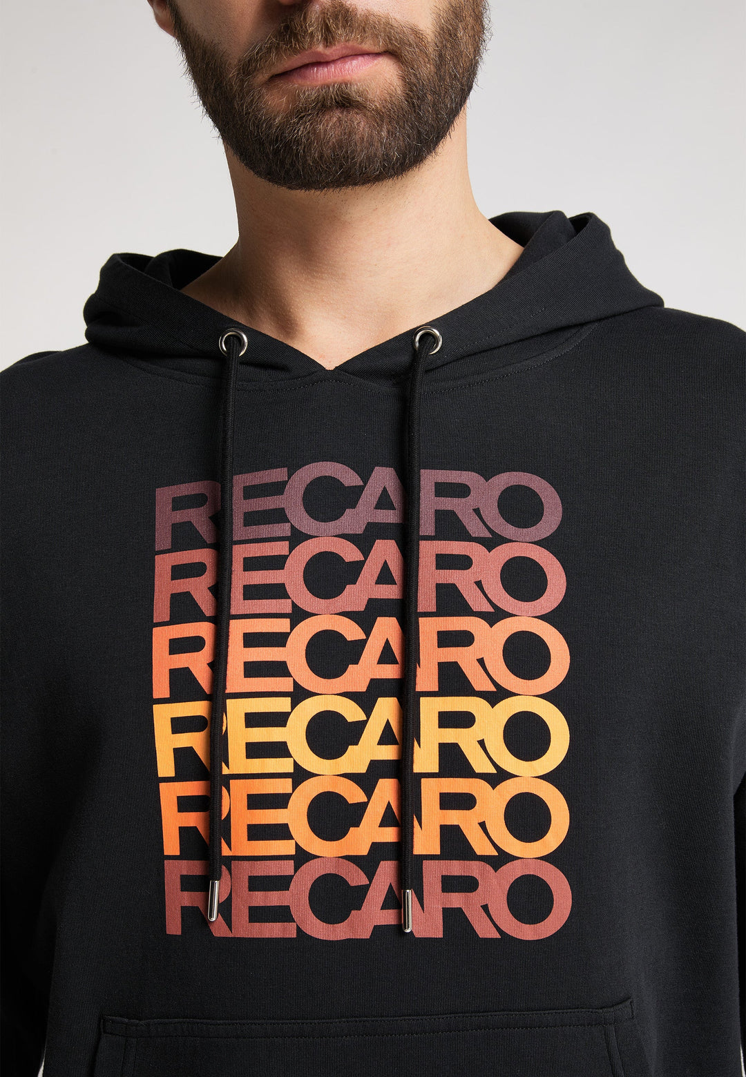 Ein Mann trägt den RECARO Hoodie Spektrum, einen schwarzen Unisex-Hoodie mit RECARO Spektrum-Stoff in einem kräftigen Rot-Gelb-Retro-Farbverlauf über der Brust. Nur die untere Hälfte seines Gesichts ist sichtbar.