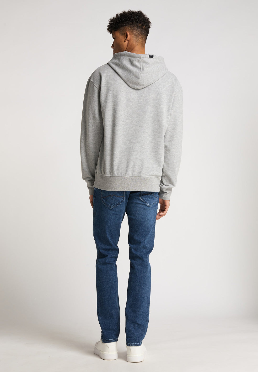Eine Person steht mit dem Rücken zur Kamera und trägt einen hellgrauen RECARO Hoodie Spektrum, Bluejeans und weiße Turnschuhe vor einem schlichten weißen Hintergrund.