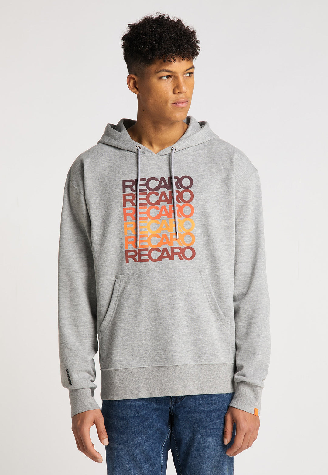 Ein junger Mann präsentiert das RECARO  Spektrum-Bundle – Hoodie & Snapback Cap – in Hellgrau mit „RECARO“ in Retro-Rot, Orange und Gelb, steht vor einem schlichten weißen Hintergrund und blickt leicht nach links.