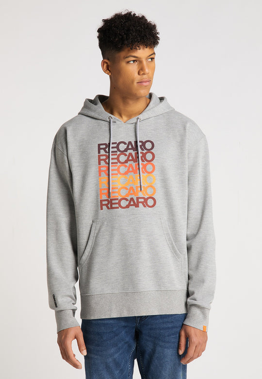 Ein junger Mann präsentiert das RECARO  Spektrum-Bundle – Hoodie & Snapback Cap – in Hellgrau mit „RECARO“ in Retro-Rot, Orange und Gelb, steht vor einem schlichten weißen Hintergrund und blickt leicht nach links.