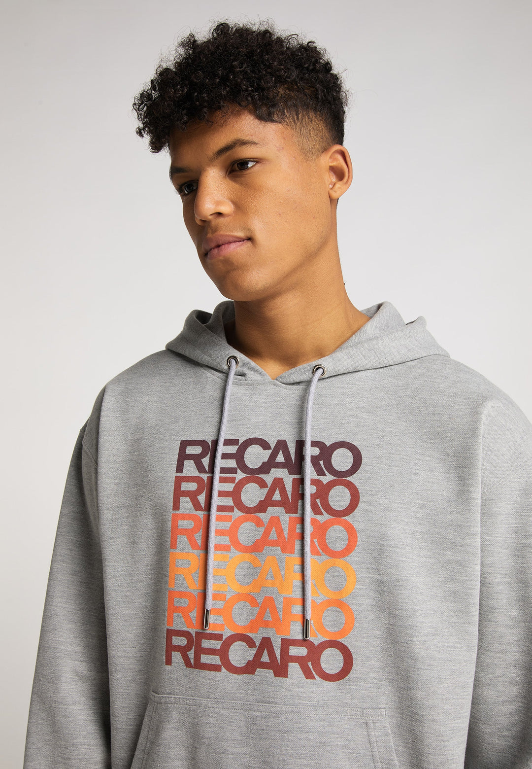 Eine junge Person mit kurzen lockigen Haaren trägt den  RECARO Hoodie Spektrum, der auf der Vorderseite RECARO in Retro-Rot, Orange und Gelb ziert. Der Hintergrund ist schlicht und hell gehalten.