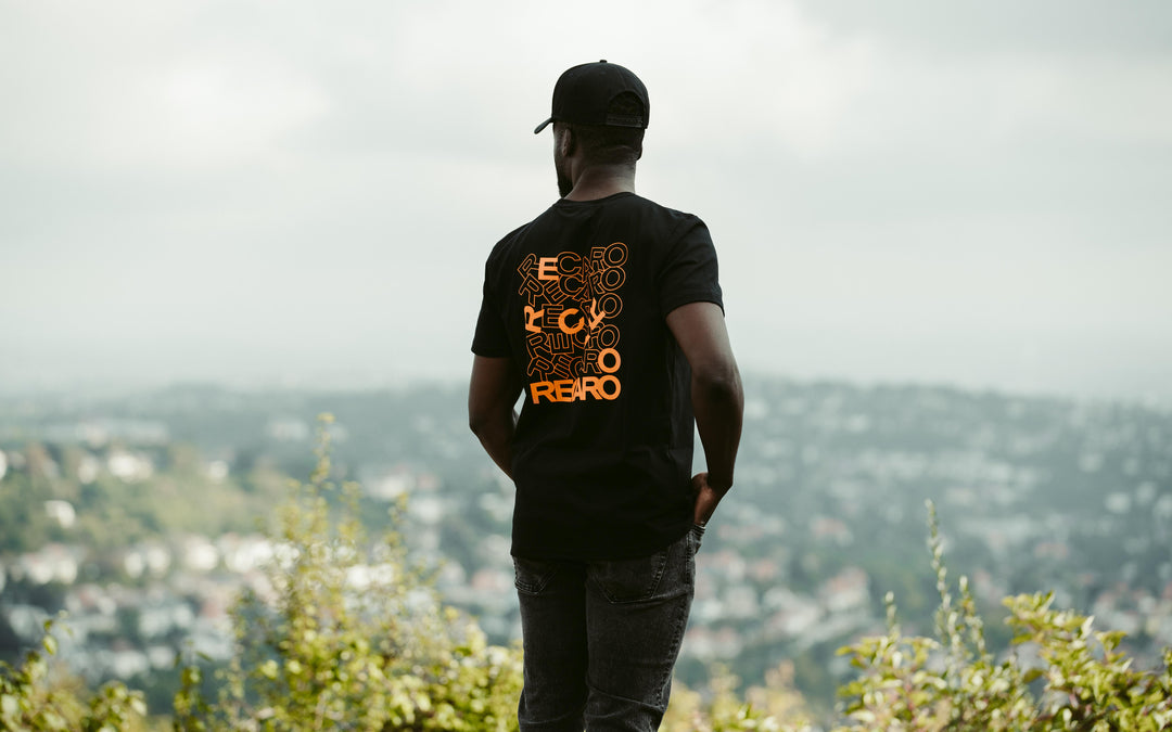 Ein Mann mit einer schwarzen Mütze und einem Recaro_Merch_Daniel T-Shirt-Rückenaufdruck steht im Freien, blickt abgewandt auf eine verschwommene Stadtlandschaft mit Grün im Vordergrund.