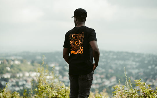 Ein Mann mit einer schwarzen Mütze und einem Recaro_Merch_Daniel T-Shirt-Rückenaufdruck steht im Freien, blickt abgewandt auf eine verschwommene Stadtlandschaft mit Grün im Vordergrund.