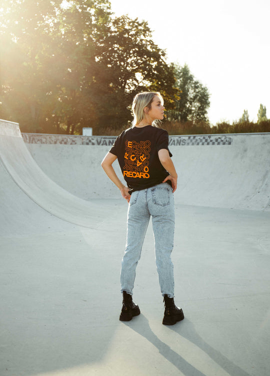 Eine blonde Frau steht mit abgewandtem Blick in einem Skatepark aus Beton. Sie trägt ein schwarzes Recaro_Merch_Daniel T-Shirt mit Rückenaufdruck, hellblaue Jeans und schwarze Stiefel. Sonnenlicht fällt durch die Bäume im Hintergrund.