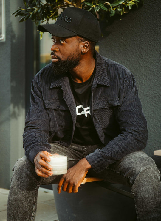 Ein Mann in einer Recaro Hemdjacke Cord Originals, schwarzer Cap und Jeans sitzt mit einem Glas Milchgetränk im Freien und blickt nachdenklich nach links. Grüne Blätter und eine graue Wand bilden den Hintergrund.