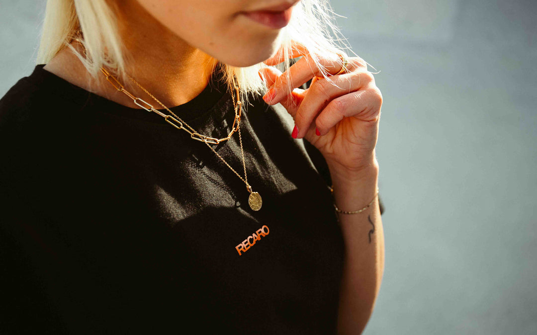 Eine Nahaufnahme einer blonden Person, die das schwarze T-Shirt mit Rückenaufdruck von RECARO trägt, dazu mehrreihige goldene Halsketten, ein goldenes Armband und roten Nagellack, und deren Haar im Sonnenlicht darauf fällt.