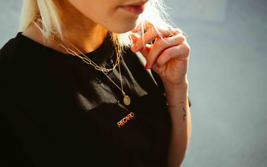 Eine Nahaufnahme einer blonden Person, die das schwarze T-Shirt mit Rückenaufdruck von RECARO trägt, dazu mehrreihige goldene Halsketten, ein goldenes Armband und roten Nagellack, und deren Haar im Sonnenlicht darauf fällt.