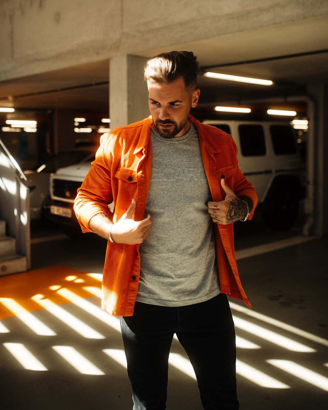 Ein Mann mit frisiertem Haar und Bart steht in einem sonnendurchfluteten Parkhaus. Er trägt eine leuchtend orange Hemdjacke Cord Originals von Recaro_Merch_Daniel über einem grauen Hemd und einer schwarzen Hose. Geometrische Schatten auf Boden und Körper erinnern an das RECARO Erbe.