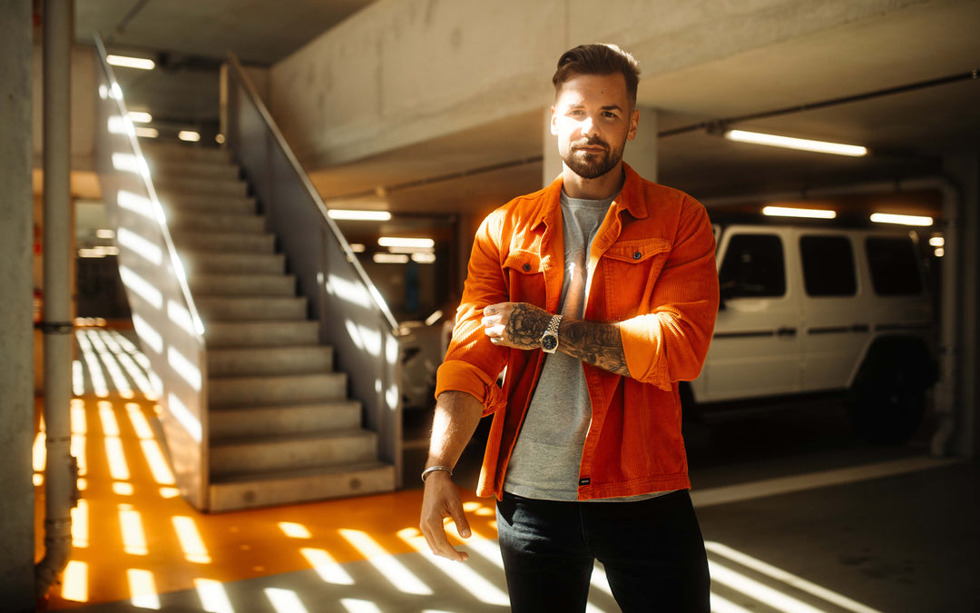 Ein tätowierter Mann in einer orangefarbenen Hemdjacke „Cord Originals“ von Recaro_Merch_Daniel und einem grauen Hemd steht in einem sonnendurchfluteten Parkhaus in der Nähe einer Treppe, wobei das gestreifte Sonnenlicht und ein weißer SUV die RECARO Heritage-Atmosphäre verstärken.