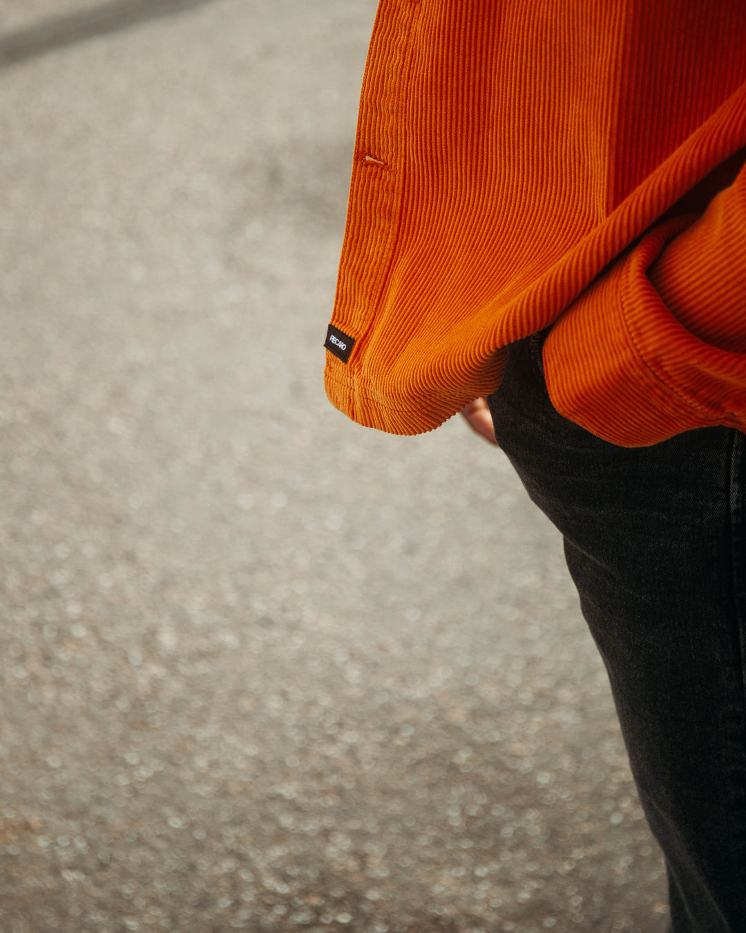 Eine Person in einer leuchtend orangefarbenen Hemdjacke „Cord Originals“ von Recaro_Merch_Daniel und dunklen Jeans steht mit einer Hand in der Tasche vor einem verschwommenen, strukturierten Bürgersteighintergrund.