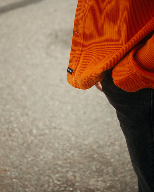 Eine Person in einer leuchtend orangefarbenen Hemdjacke „Cord Originals“ von Recaro_Merch_Daniel und dunklen Jeans steht mit einer Hand in der Tasche vor einem verschwommenen, strukturierten Bürgersteighintergrund.