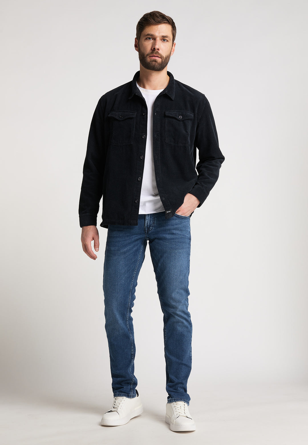 Ein bärtiger Mann blickt nach vorne und trägt eine schwarze Recaro_Merch_Daniel Hemdjacke Cord Originals über einem weißen T-Shirt, Bluejeans und weißen Turnschuhen vor einem schlichten hellen Hintergrund.