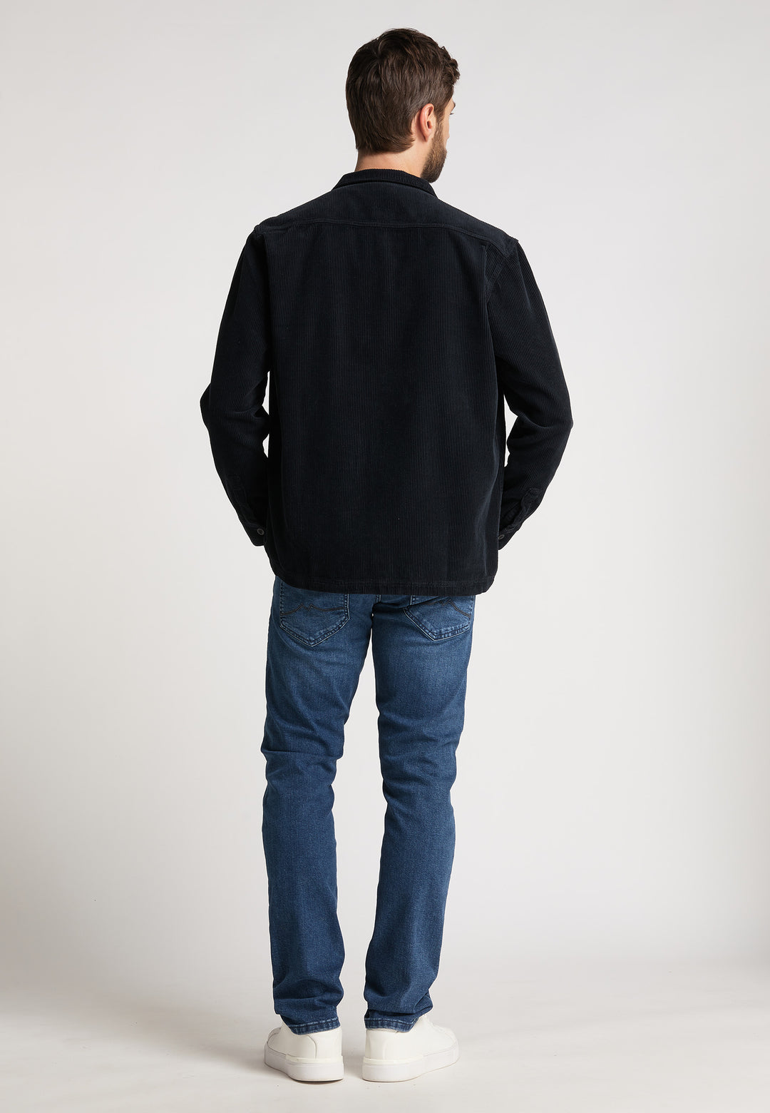 Ein Mann steht mit dem Rücken zur Kamera und trägt eine schwarze Recaro_Merch_Daniel Hemdjacke Cord Originals, Bluejeans und weiße Turnschuhe vor einem schlichten hellen Hintergrund.